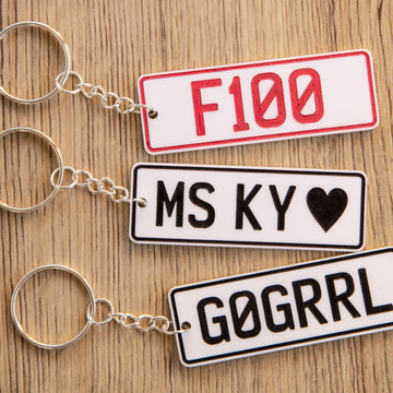 Three Number Plate Personalised Key Rings rest on a wooden surface. Each custom acrylic keyring features unique text—F100 in red, MS KY ♥, and G0GRRL in black—ideal gifts for car enthusiasts.