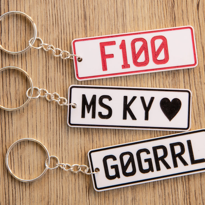 Three Number Plate Personalised Key Rings rest on a wooden surface. Each custom acrylic keyring features unique text—F100 in red, MS KY ♥, and G0GRRL in black—ideal gifts for car enthusiasts.
