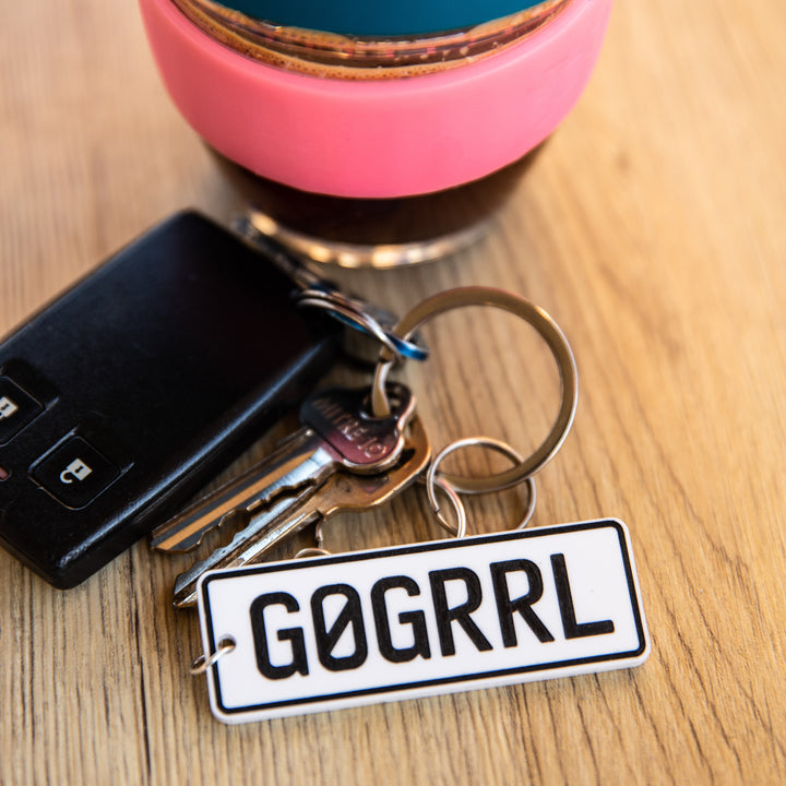 A Number Plate Personalised Key Ring with "GØGRRL" rests beside a reusable cup with a pink band on a wooden surface—an ideal gift for any car enthusiast.