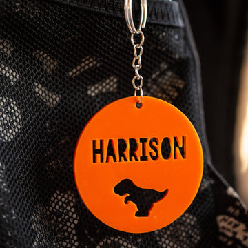An Icon Personalised Bag Tag in orange acrylic, circular with a dinosaur silhouette and the name HARRISON cut out, hangs from a metal keyring on black mesh fabric.
