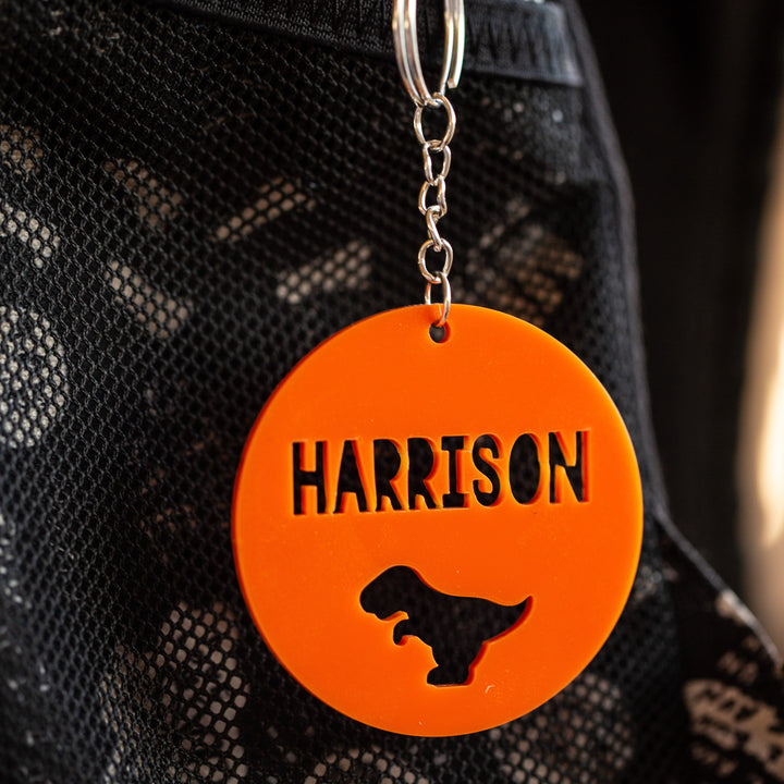 An Icon Personalised Bag Tag in orange acrylic, circular with a dinosaur silhouette and the name HARRISON cut out, hangs from a metal keyring on black mesh fabric.