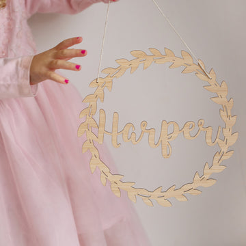 A child in a pink dress with matching nail polish holds the Personalised Flora Name Wreath—bamboo wall art featuring "Harper" in cursive. Set against a plain, light background, it’s an ideal nursery decor piece.