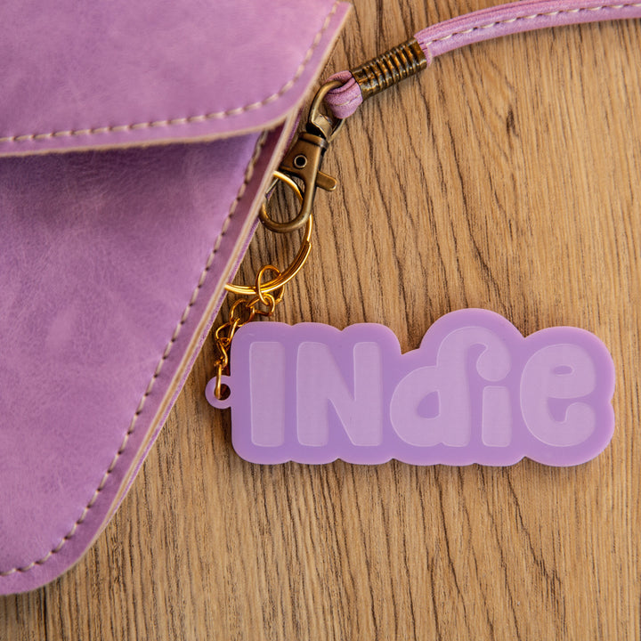 A close-up of a lavender purse with a Bubblegum Personalised Bag Tag featuring "Indie" in bold pastel purple letters, resting on a light wooden surface.