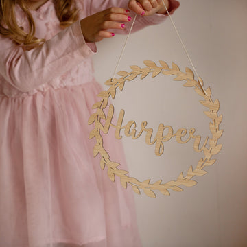 A young girl in a pink dress holds the Personalised Flora Name Wreath, featuring "Harper" in cursive. Her pink-painted nails stand out as she displays this charming bamboo ornament, perfect as adorable nursery decor against a light background.