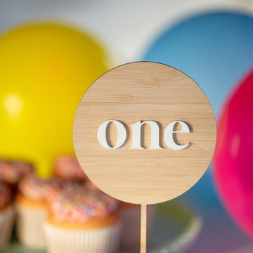 A wooden sign reading "one" stands before colorful yellow, blue, and pink balloons, with cupcakes topped with sprinkles and a Simple Personalised Cake Topper in the background.