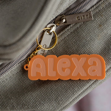 A Bubblegum Personalised Bag Tag in orange with "Alexa" in bubble letters is attached to the gray zipper of a fabric bag.