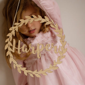 A young girl in a pink dress holds the Personalised Flora Name Wreath—bamboo wall art featuring leaf details and a custom name in cursive—partially covering her face, making it a charming touch for nursery decor.