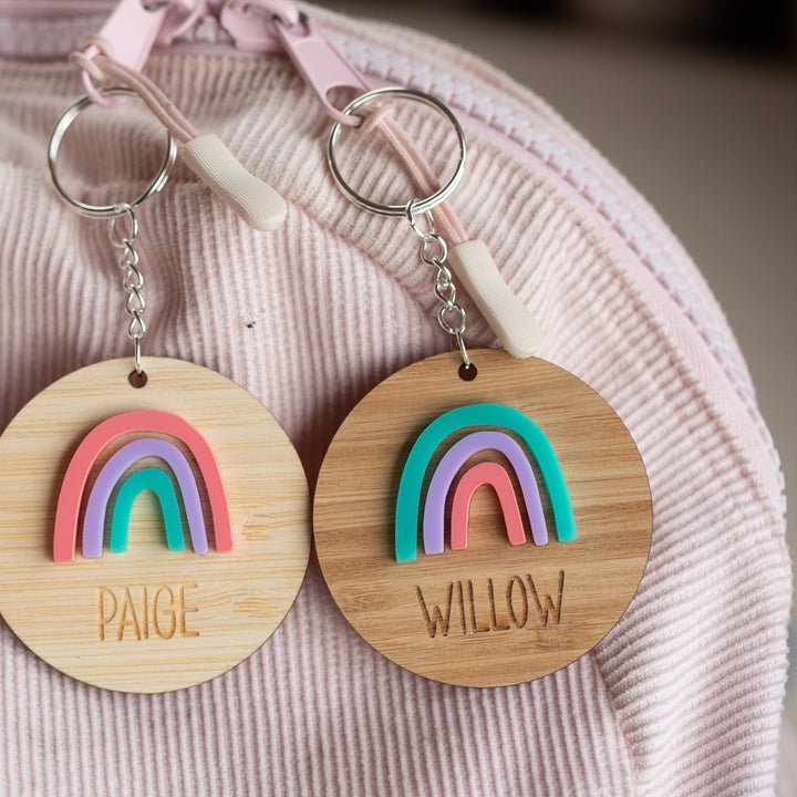 Two round wooden Rainbow Personalised Bag Tags, featuring colorful designs and engraved names “PAIGE” and “WILLOW,” hang from a pink-striped bag—perfect for adding a unique, fun touch to any backpack or schoolbag.