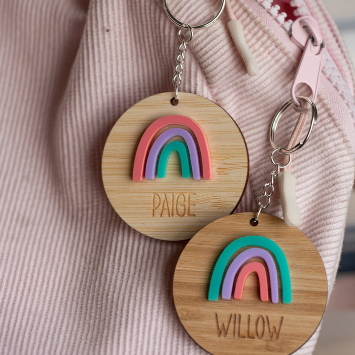 Two round wooden Rainbow Personalised Bag Tags with colorful designs and engraved names PAIGE and WILLOW, attached to a pink corduroy bag with a light pink zipper—perfect for kids.