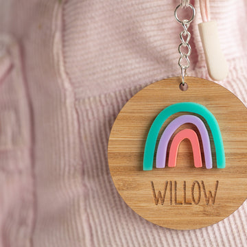 The Rainbow Personalised Bag Tag features a wooden rainbow design in teal, purple, and pink with the name WILLOW engraved below. Attached to a pink corduroy bag or jacket, it adds individuality and charm to any accessory.