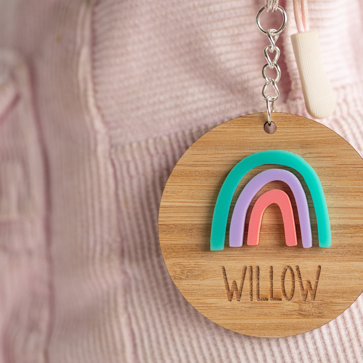 The Rainbow Personalised Bag Tag features a wooden rainbow design in teal, purple, and pink with the name WILLOW engraved below. Attached to a pink corduroy bag or jacket, it adds individuality and charm to any accessory.