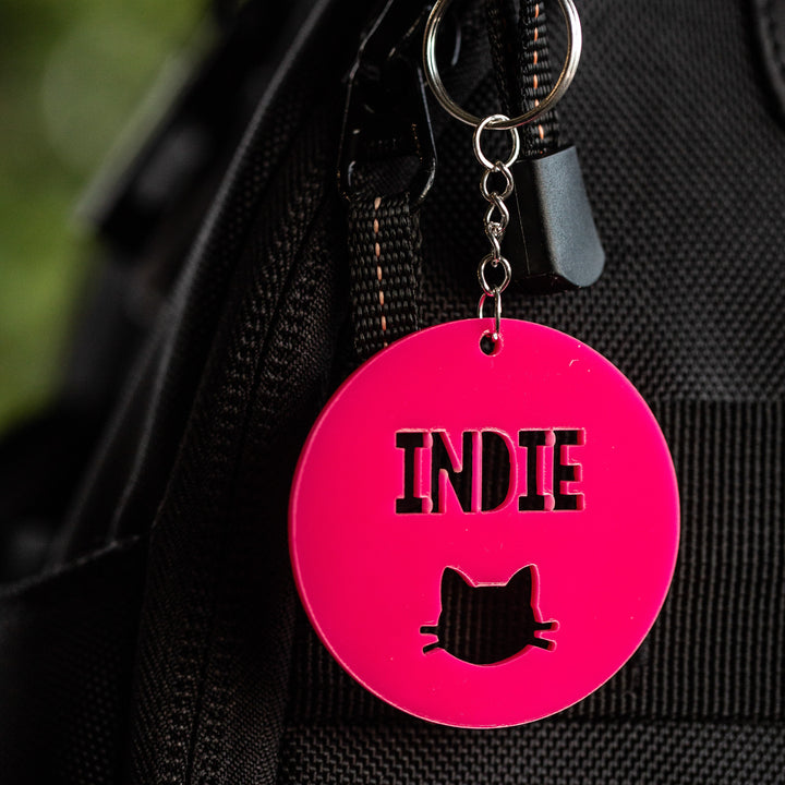 A vibrant pink circular Icon Personalised Bag Tag featuring the word INDIE and a cat head silhouette cut-out, shown attached to a black bag.