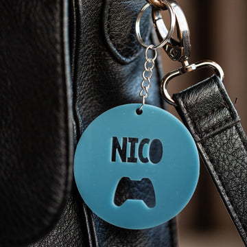The Icon Personalised Bag Tag, featuring a blue round acrylic design with the name NICO and a game controller cutout, hangs from a metal keyring on a black leather bag.