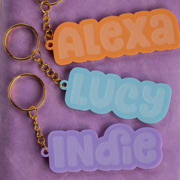 Three Bubblegum Personalised Bag Tags with gold rings lie on a purple surface, each showcasing a name—ALEXA in orange, LUCY in blue, and INDIE in lavender—in bold, playful letters for a fun custom accessory.