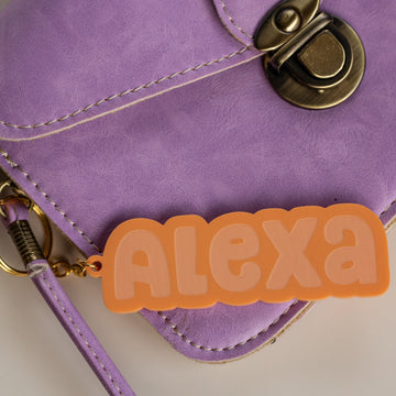 A close-up of a purple heart-shaped bag with a brass clasp, featuring the Bubblegum Personalised Bag Tag—a peach acrylic keyring with “ALEXA” in bold, rounded letters.