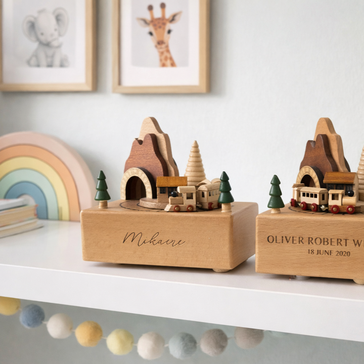 Two personalised wooden music boxes with trains and trees sit on a white shelf. One is labeled "Personalised Wooden Music Box." Behind them are a rainbow ornament, children&