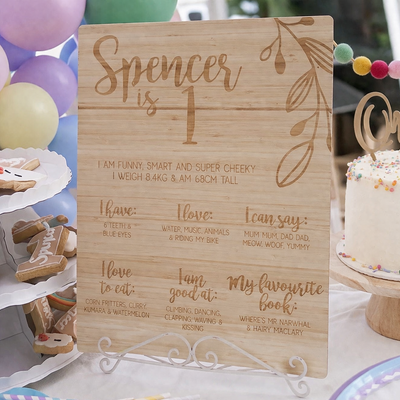 Wooden sign with engraved birthday information for a child, surrounded by party decorations including a cake and balloons.
