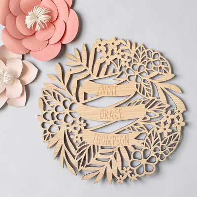 The Personalised Tropics Wall Plaque is a round wooden name plaque featuring "Indie Grace Thompson" engraved in the center, surrounded by detailed flowers and leaves, with pink and white paper flowers on the upper left for a tropical touch.