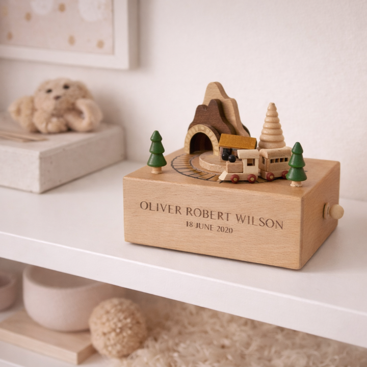 A Personalised Wooden Music Box featuring miniature trees, a mountain, and a train is displayed on a white shelf. Engraved with "Oliver Robert Wilson, 18 June 2020," it&