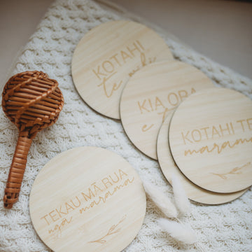 The Te Reo Māori Baby Milestones Set (available in 2 set sizes) includes a woven rattle and four eco-friendly bamboo milestone cards with Te Reo Māori words, displayed on a cream knitted blanket with two dried white flowers.