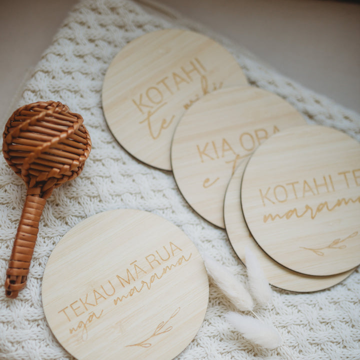 The Te Reo Māori Baby Milestones Set (available in 2 set sizes) includes a woven rattle and four eco-friendly bamboo milestone cards with Te Reo Māori words, displayed on a cream knitted blanket with two dried white flowers.
