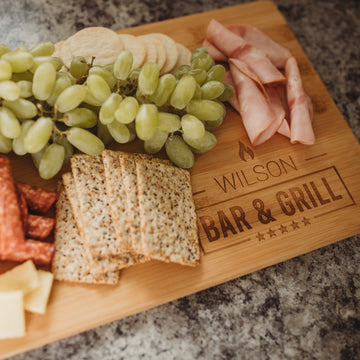 A Personalised Bamboo Platter/Serve Board (Two Sizes Available) engraved with Wilson Bar & Grill displays green grapes, crackers, rolled ham slices, salami, and cheese cubes—a perfect gift that New Zealand food lovers will cherish.