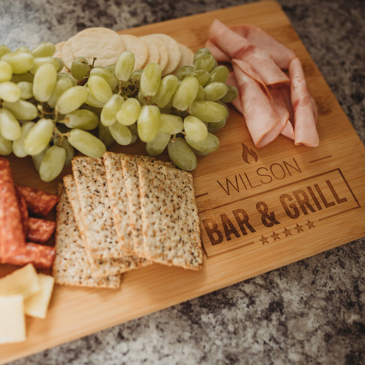 A Personalised Bamboo Platter/Serve Board (Two Sizes Available) engraved with Wilson Bar & Grill displays green grapes, crackers, rolled ham slices, salami, and cheese cubes—a perfect gift that New Zealand food lovers will cherish.