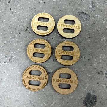 Six round wooden Removable PomPom Buttons with two holes each are displayed in two columns on a grey speckled surface, three read "HANDMADE" for crochet projects, and three say "REMOVABLE."