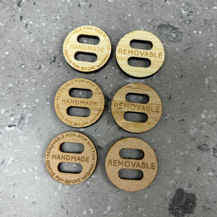 Six round wooden Removable PomPom Buttons with two holes each are displayed in two columns on a grey speckled surface, three read "HANDMADE" for crochet projects, and three say "REMOVABLE."