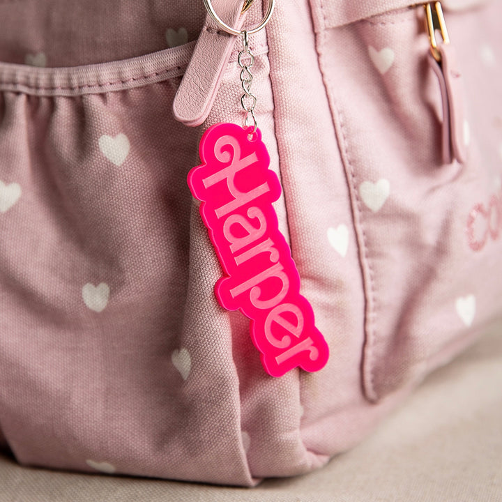 A pink bag with white heart patterns features a Barbie-inspired Personalised Bag Tag as a keychain displaying "Harper" in bold pink letters hanging from the zipper pull.