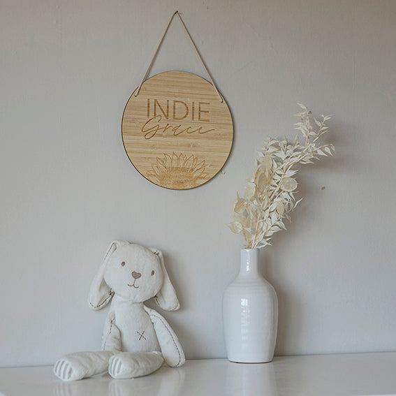 A soft white stuffed bunny sits beside a white vase with dried pale foliage on a white surface. Above them, a round Personalised Sunflower Plaque hangs on the wall.