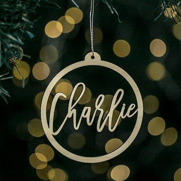 A round bamboo Halo Christmas Bauble featuring the name Charlie in script hangs on a tree with blurred yellow lights—a charming personalised keepsake, gift pouch included.