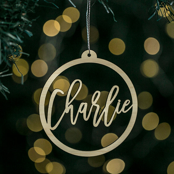 A round bamboo Halo Christmas Bauble featuring the name Charlie in script hangs on a tree with blurred yellow lights—a charming personalised keepsake, gift pouch included.