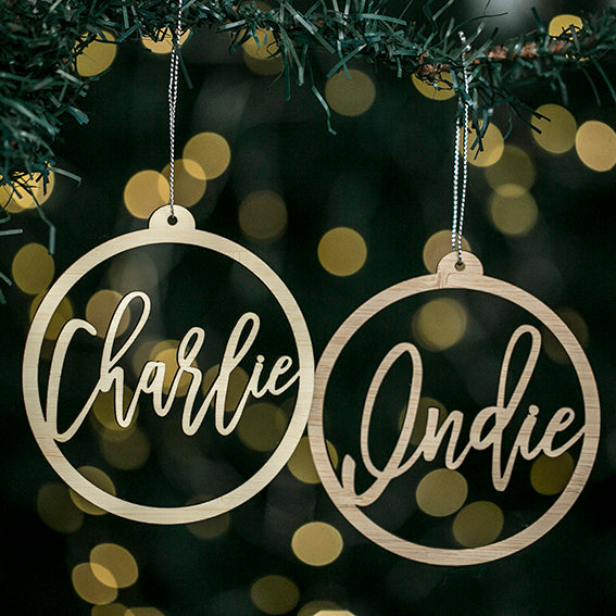 Two Halo Christmas Bauble ornaments hang from a tree branch, each with a circular frame and cursive names—one reads Charlie, the other Indie—against blurred yellow lights. A Personalised keepsake gift pouch is included.