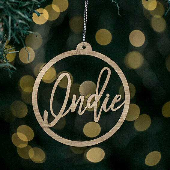A bamboo Halo Christmas Bauble with "Indie" in cursive hangs from a tree branch amid festive yellow lights. It comes in a keepsake pouch—perfect for gifting or treasuring as a personalised ornament.