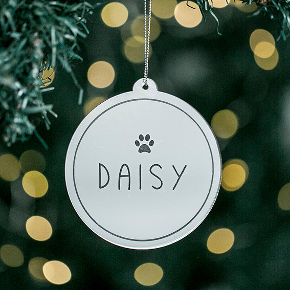 An Etched Pets Christmas Bauble, engraved with a paw print and the name DAISY, hangs from a tree branch amid softly blurred warm lights in the background.