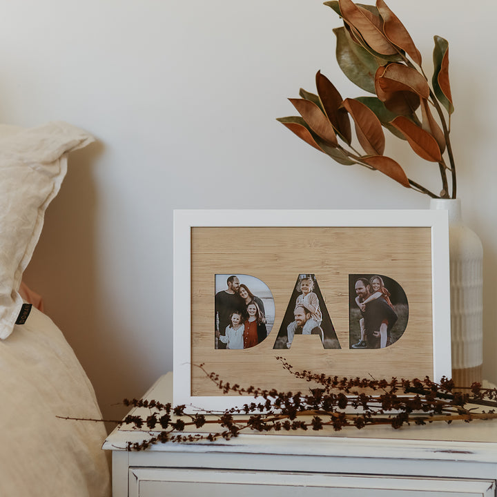 A DAD photo frame insert (frame not included) with family photos in the letters sits on a white bedside table next to a vase of dried leaves—an ideal New Zealand gift. Part of a bed with beige linens is visible on the left.
