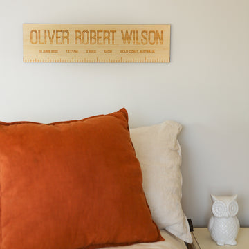 A Personalised Birth Ruler featuring birth details hangs on a light wall above a bed with rust and beige pillows. A white ceramic owl sits on the bedside table, adding charm to this cosy space.
