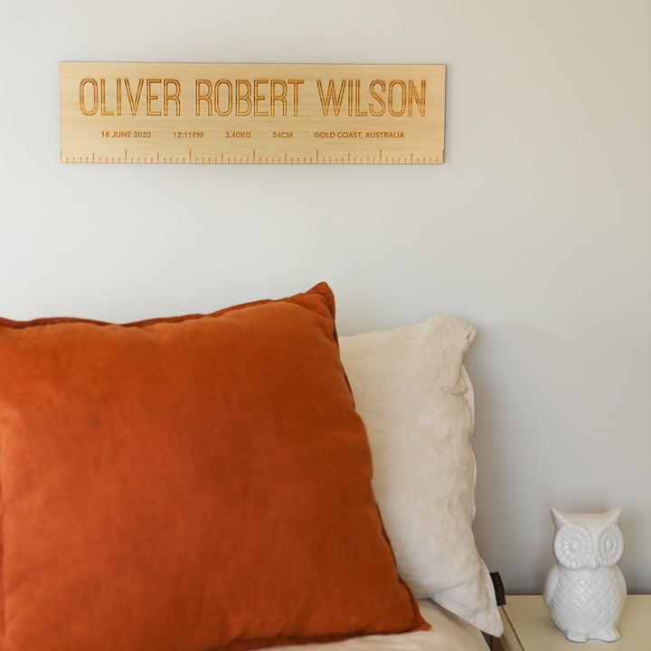 A Personalised Birth Ruler featuring birth details hangs on a light wall above a bed with rust and beige pillows. A white ceramic owl sits on the bedside table, adding charm to this cosy space.
