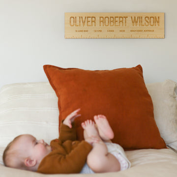 A baby in brown clothing lies on a white couch with a rust pillow behind them and a Personalised Birth Ruler on the wall displaying "Oliver Robert Wilson" along with his birth details and measurements.