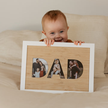 A smiling baby sits on a bed holding a DAD Photo Frame Insert (frame not included), which displays family photos inside the letters. Made with a bamboo A4 insert, it&
