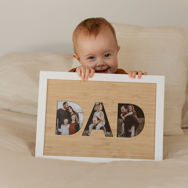 A smiling baby sits on a bed holding a DAD Photo Frame Insert (frame not included), which displays family photos inside the letters. Made with a bamboo A4 insert, it&