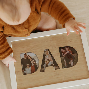 A child in a brown outfit holds a DAD Photo Frame Insert (frame not included)—each letter displays cherished family moments, making it a perfect New Zealand gift. The A4 bamboo insert beautifully celebrates special memories together.