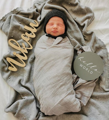 A wooden wall name saying Nikau, sitting next to a new born baby and a sign saying Hello World.