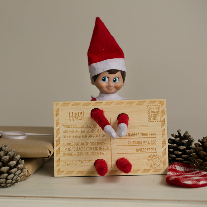 The Elf on the Shelf Arrival Postcard features a Christmas elf doll behind a personalised postcard with an engraved message, set among pinecones, a wrapped gift, and red plaid on a wooden surface for a festive touch.