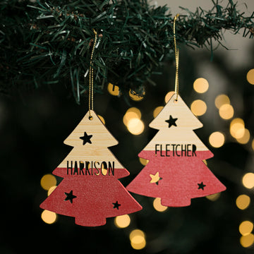 Two wooden personalised ornaments with cut-out stars hang on the Evergreen Christmas Tree. One is labeled “HARRISON,” the other “FLETCHER,” while warm, blurred lights glow softly in the background.