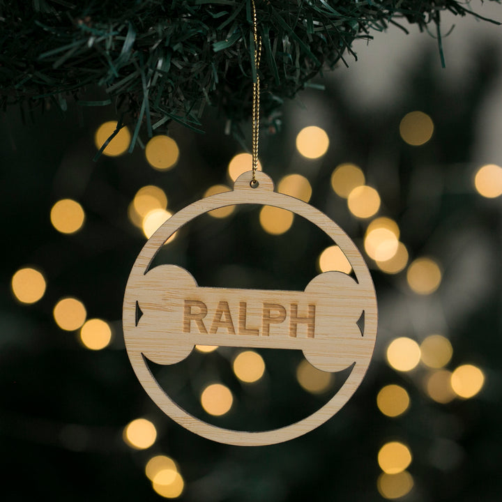 The Dog Bone Christmas Bauble, a circular bamboo ornament engraved with RALPH, hangs from a tree branch as blurred yellow lights twinkle in the background—an eco-friendly personalised decoration for your Christmas tree.