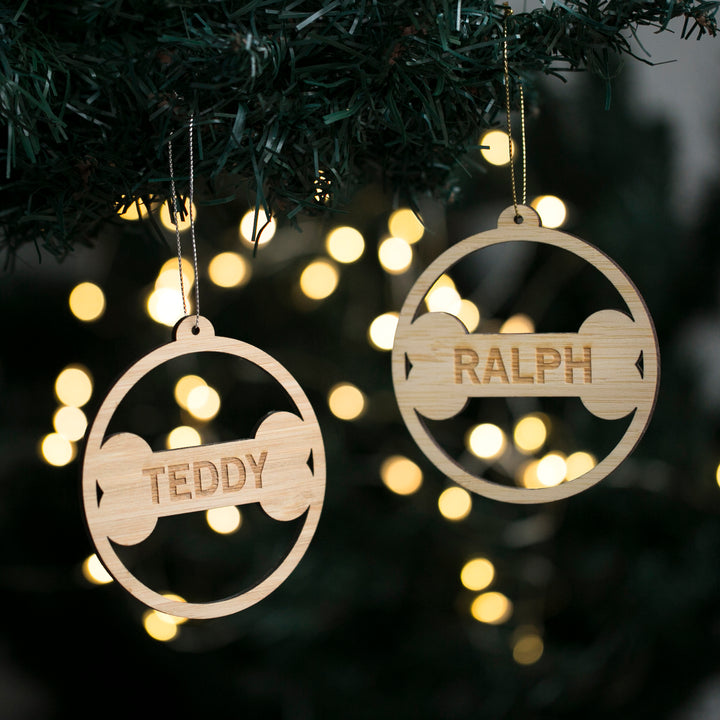 Two Dog Bone Christmas Bauble ornaments made from eco-friendly bamboo hang on a tree. Each circular bauble features a personalized dog bone engraved with the names Teddy and Ralph, with softly glowing string lights in the background.
