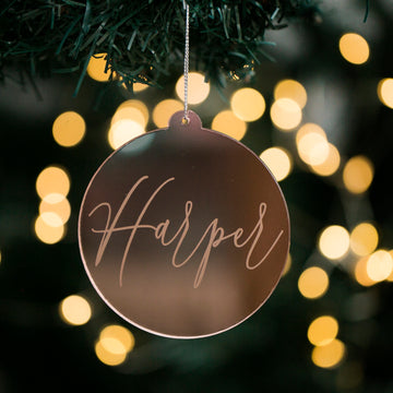 The Script Christmas Bauble, a round mirrored acrylic ornament with "Harper" elegantly scripted on it, hangs from a tree amid glowing holiday lights—making it the perfect personalised decoration for the festive season.