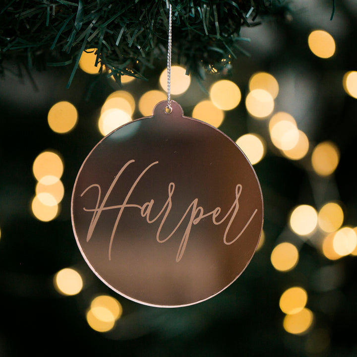 The Script Christmas Bauble, a round mirrored acrylic ornament with "Harper" elegantly scripted on it, hangs from a tree amid glowing holiday lights—making it the perfect personalised decoration for the festive season.